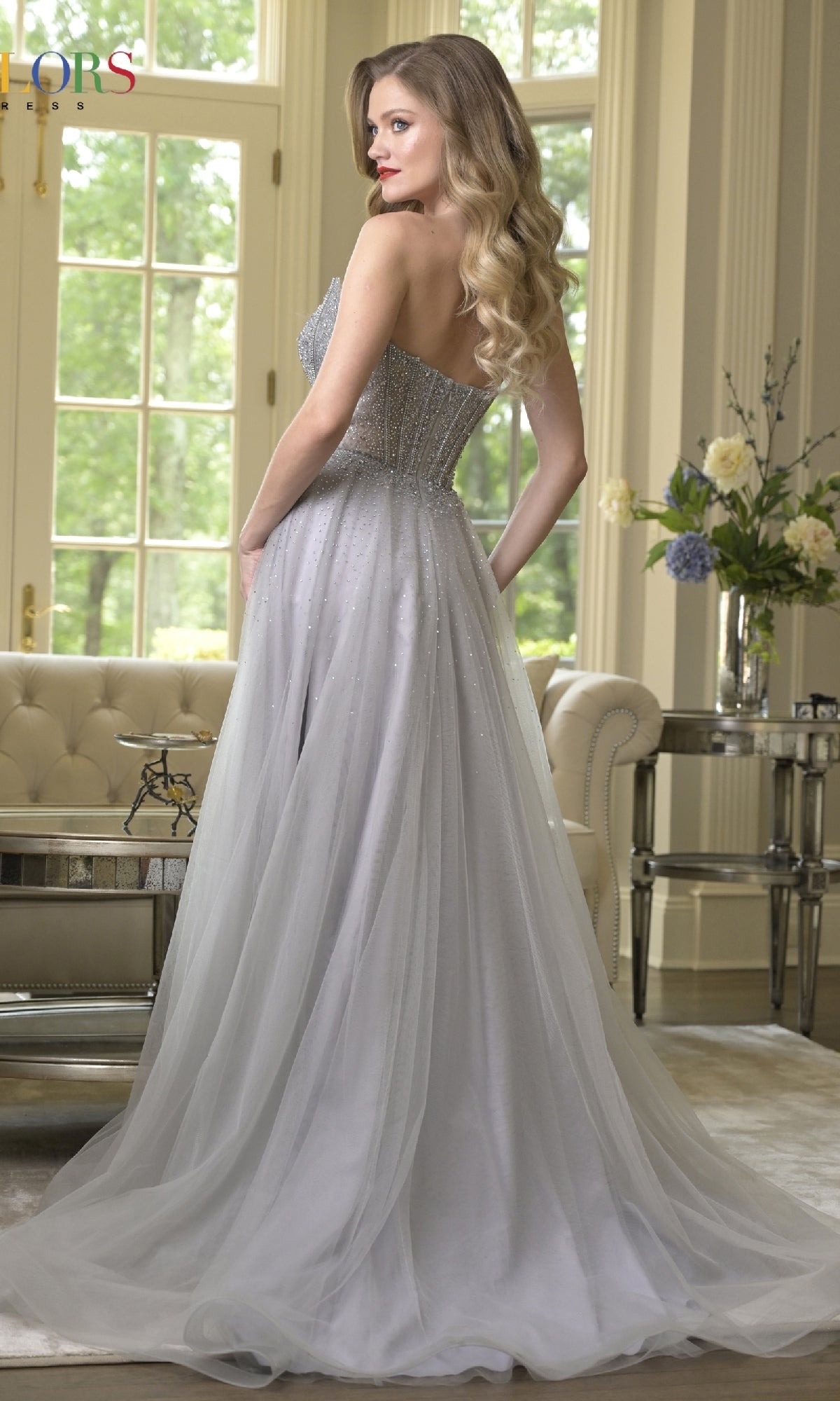 A woman with long blonde hair models the Colors Dress 3879, a strapless prom dress with an embellished corset bodice and tulle A-line skirt, standing in an elegant room with large windows, a sofa, and flowers on a side table.